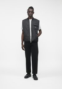 Αμάνικο μαύρο γιλέκο με ραφές, με μπροστινή τσέπη, φορεμένο πάνω από γκρι μπλούζα και συνδυασμένο με μαύρο παντελόνι και μαύρα παπούτσια. Λείο ύφασμα.