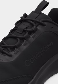 Gros plan sur une basket noire Calvin Klein avec des lacets épais, une languette de tirage et une semelle texturée, montrant le nom de la marque embossé sur le côté.