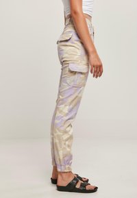Beige og lavendel camo cargo bukser med elastiske mansjetter, sidelommer og løs passform, laget av slitesterkt stoff. Stilt med svarte sandaler.