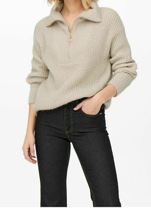 Beige knitted sweater with a half-zip collar, textured pattern, and ribbed cuffs, paired with dark jeans featuring yellow stitching.
