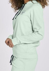 Sudadera verde menta con un bolsillo frontal, capucha con cordones y acentos en azul marino. Hecha de una tela suave, con puños y dobladillo de canalé.