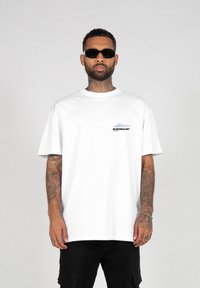 Hvit bomull T-skjorte med kort erm, rund hals, med en blå og svart logo på venstre brystområde. Brukt sammen med svarte cargo-shorts.