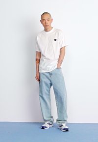 White short-sleeve t-shirt with a small black heart logo, light blue baggy jeans, and white sneakers with blue accents.
