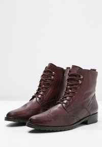 Botas de tobillo de cuero marrón con cordones en la parte delantera, detalles de brogue, tacón bajo y cremallera lateral para facilitar el uso. Textura suave con un acabado pulido.