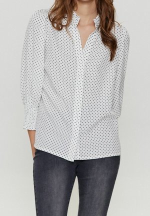 Femme portant un chemisier blanc à pois noirs avec des manches trois-quarts bouffantes, associé à un jean foncé.