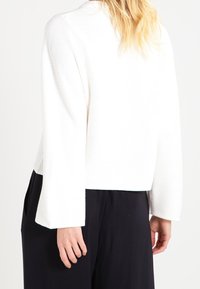 Vitt kortat tröja med ribbad textur, breda trumpetärmar och rak nederkant, bärs över svarta byxor. Bakifrån visas halsringningen och ärmdesignen.