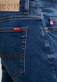 Denim jeans in dark blue with a smooth texture. Features red branded tags, contrast stitching, and a zigzag pattern on the back pocket.