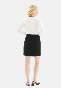 Jupe noire mi-longue en tissu texturé avec une taille. Associée à un haut à manches longues de couleur claire et des chaussures à talons noires.