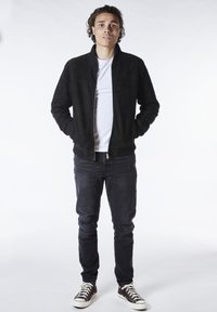 Svart mockabomberjacka, vit t-shirt under, slitna mörka jeans och mörka högskaftade sneakers. Enkelt, åtsittande design med dragkedja.