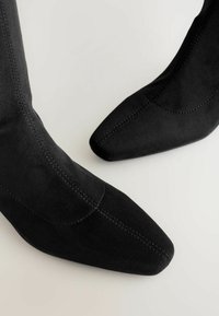 Bottines noires en daim avec un embout pointu, dotées de coutures apparentes et d'une texture lisse. Aucun matériel ni ornement présent.