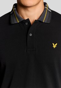 Μαύρο πόλο πουκάμισο με υφασμένο γιακά που έχει γκρι και κίτρινες ρίγες, δύο κουμπιά στη λαιμόκοψη και ένα μικρό κίτρινο λογότυπο αετού στο στήθος.