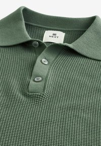Grøn tekstureret polo shirt med krave, tre knaplukninger og mærkeetiket. Fremstillet af et strikstof med et subtilt mønster.