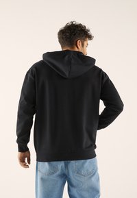 Schwarzer Pullover-Hoodie mit verstellbarem Kapuzenzug, gerippten Bündchen und Saum, kombiniert mit hellblauen Jeans, von hinten betrachtet. Glatte Stoffstruktur.