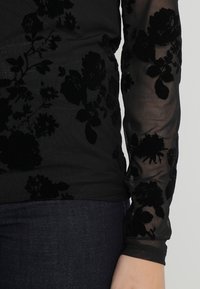 Svart transparent topp med långa ärmar, prydd med ett strukturerat blommigt sammetmönster. Buren över mörk denim, vilket framhäver den delikata designen.