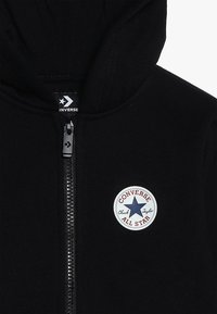 Schwarzer Hoodie mit durchgehendem Reißverschluss. Verfügt über einen runden weißen Aufnäher mit einem blauen Stern und roter Schrift, die "CONVERSE CHUCK TAYLOR ALL STAR" lautet.