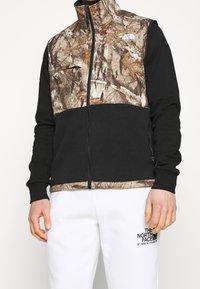 Kurtka polarowa z czarną dolną częścią i wielotonową kamuflażową górą. Posiada wysoki kołnierz i kieszenie na zamek.