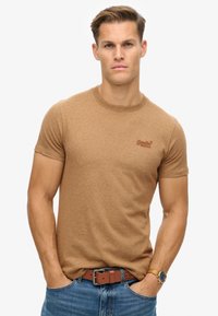 T-shirt corta marrone con collo rotondo e un piccolo logo sul petto, abbinata a jeans blu e una cintura marrone. Texture liscia.