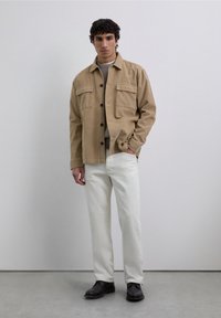 Hombre de pie en interiores que lleva puesta una chaqueta de pana beige, un suéter gris, pantalones blancos y zapatos negros, contra un fondo gris liso.