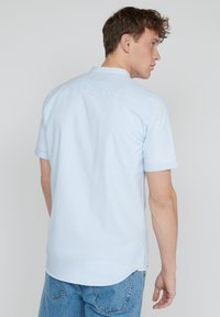 Man met krullend haar die een lichtblauw gestreept shirt met korte mouwen en blauwe denim jeans draagt, met de rug naar de camera gekeerd tegen een effen achtergrond.