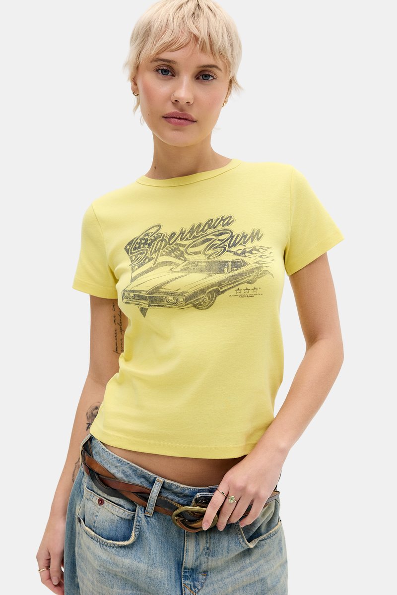 Νεαρό άτομο με κοντά ξανθά μαλλιά φοράει κίτρινο T-shirt με σχέδιο "Supernova Burn" και φαρδιά μπλε τζιν με καφέ ζώνη σε λευκό φόντο.