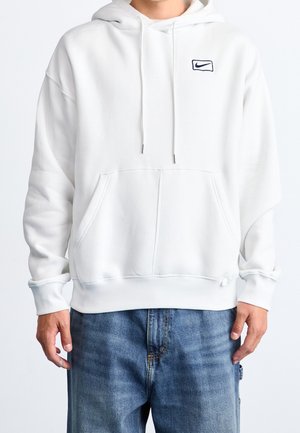 Mężczyzna w białej bluzie Nike z kapturem i przednią kieszenią oraz jasnoniebieskich dżinsach, stojący na tle jednolitego jasnego tła.