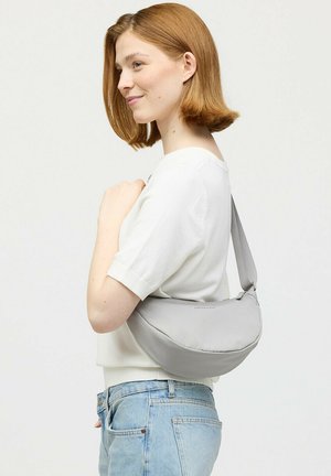 Young woman with short red hair wearing a white top and light blue jeans carrying a grey shoulder bag across her back.