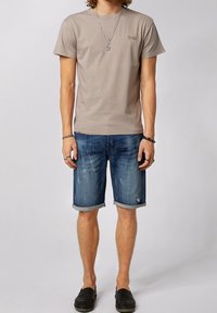 T-shirt in cotone beige chiaro con maniche corte, abbinata a pantaloni shorts in denim blu con orlo arrotolato e dettagli strappati, completata con scarpe slip-on nere.