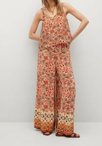 Kombinezon w kwiatowy wzór z szerokimi nogawkami, wykonany z lekkiego materiału. Posiada cienkie ramiączka i luźny krój w ciepłych odcieniach czerwieni i beżu.
