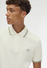 Camisa polo blanca con cuello a rayas verde oliva, dos botones y logo en el pecho. Tejido texturizado y diseño ajustado.