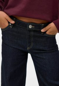 Jean bleu foncé en denim, présentant un design classique à cinq poches, une fermeture à bouton argenté et des coutures jaunes contrastantes.