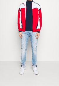 Röd- och vit träningsjacka med hög krage, marinblå polotröja, ljusblå jeans och vita sneakers. Jackan har kontrasterande paneler.