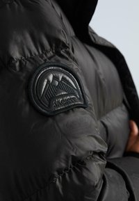 Melnā pūkainā jaka ar spīdīgu apdari. Iezīmēta ar teksturētu melnu plāksteri ar kalnu dizainu un "JAKA" tekstu kreisajā piedurknē.