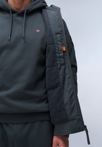 Giacca piumino grigia scura con un morbido rivestimento interno in fleece nero, dotata di zip laterale e dettagli arancioni. Il cappuccio sottostante ha coulisse regolabili.