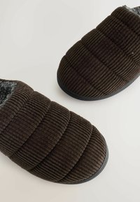Hnedé kordurové papuče s polstrovaným, textúrovaným povrchom a čiernou gumovou podrážkou. Majú mäkkú podšívku a zaoblený tvar špičky.
