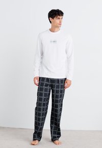 Cămașă albă cu mâneci lungi, decorată cu un grafic în dungi orizontale; asortată cu pantaloni negri în carouri, cu linii albe de accent și croială lejeră.