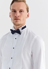 Biała koszula z przodu z wyszywanymi fałdami, czarnymi guzikami i granatowym krawatem w muszkę. Gładki materiał, klasyczny kołnierzyk i dopasowany krój.