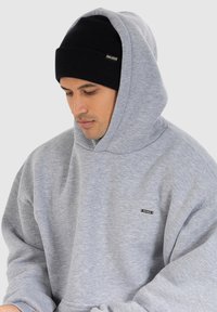 Felpa grigia in cotone con un grande cappuccio, polsini a coste e una piccola etichetta nera. Indossata con un berretto a maglia nero aderente.