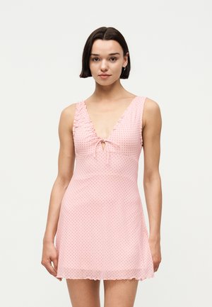 Vrouw met kort donker haar die een lichtroze mouwloze jurk draagt met kleine zwarte stippen en een strik in het midden van de halslijn, staande tegen een witte achtergrond.