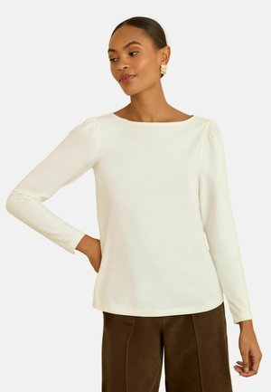 Weiße Langarmbluse mit rundem Halsausschnitt und leicht gepufften Ärmeln, gefertigt aus einem glatten Stoff. Kombiniert mit braunen, texturierten Hosen.