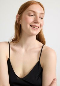 Svart satin camisole med tunna axelband och V-ringning, med en slät yta och en lätt glans. Modellen har långt hår och ljus hy.