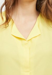 Žlutá blůza s výstřihem do V, zapínáním na knoflíky a hladkým, lehkým materiálem. Má jemný lesk a volný střih.