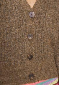 Lähikuva ruskeasta neulotusta neuletakista, jossa on neljä suurta nappia värikkään raidallisen vaatteen päällä.