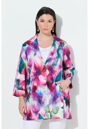 Kvinde iført en farverig blomsterblazer med pink, lilla og grønne nuancer over en hvid top, poserer med den ene hånd i lommen foran en hvid baggrund.