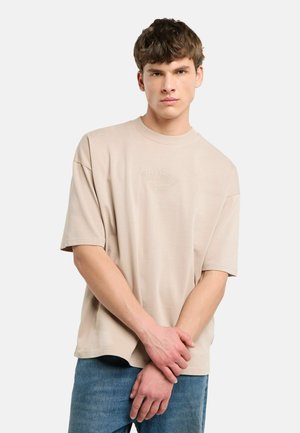 Junger Mann mit kurzen braunen Haaren, der ein beige oversized T-Shirt und blaue Jeans trägt und mit verschränkten Händen vor dem Körper steht.