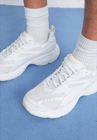 White athletic shoes featuring a mesh upper, cushioned sole, and textured accents. Worn with ribbed white socks on a blue surface.