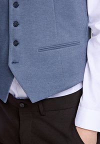 Blå mönstrad väst med fem svarta knappar, en ficka på vänster sida, ihop med en vit skjorta och svarta byxor. Texturerad tyg.