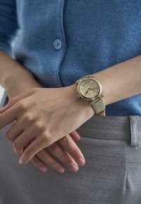 Auksinio tono laikrodis su tekstūruota smėlio spalvos apyranke. Apvali ciferblato dalis pasižymi šviesia rodykle ir linijiniais žymekliais. Dėvimas ant neutraalaus odos tono rankos.