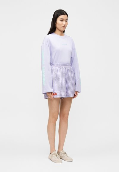 Γυναίκα που στέκεται φορώντας ένα ανοιχτό μωβ μακρυμάνικο πουκάμισο με κείμενο στο μανίκι και μια μωβ φούστα με καρό σχέδιο, μπεζ αθλητικά παπούτσια.