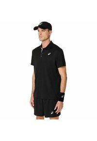 Uomo con berretto sportivo nero, polo, pantaloni corti e braccialetto con loghi ASICS bianchi, in piedi di fronte a uno sfondo bianco.