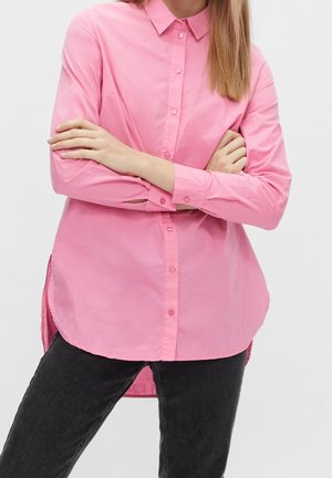 Personne portant une chemise rose à manches longues avec col et pantalon noir, bras croisés sur la poitrine devant un fond uni.